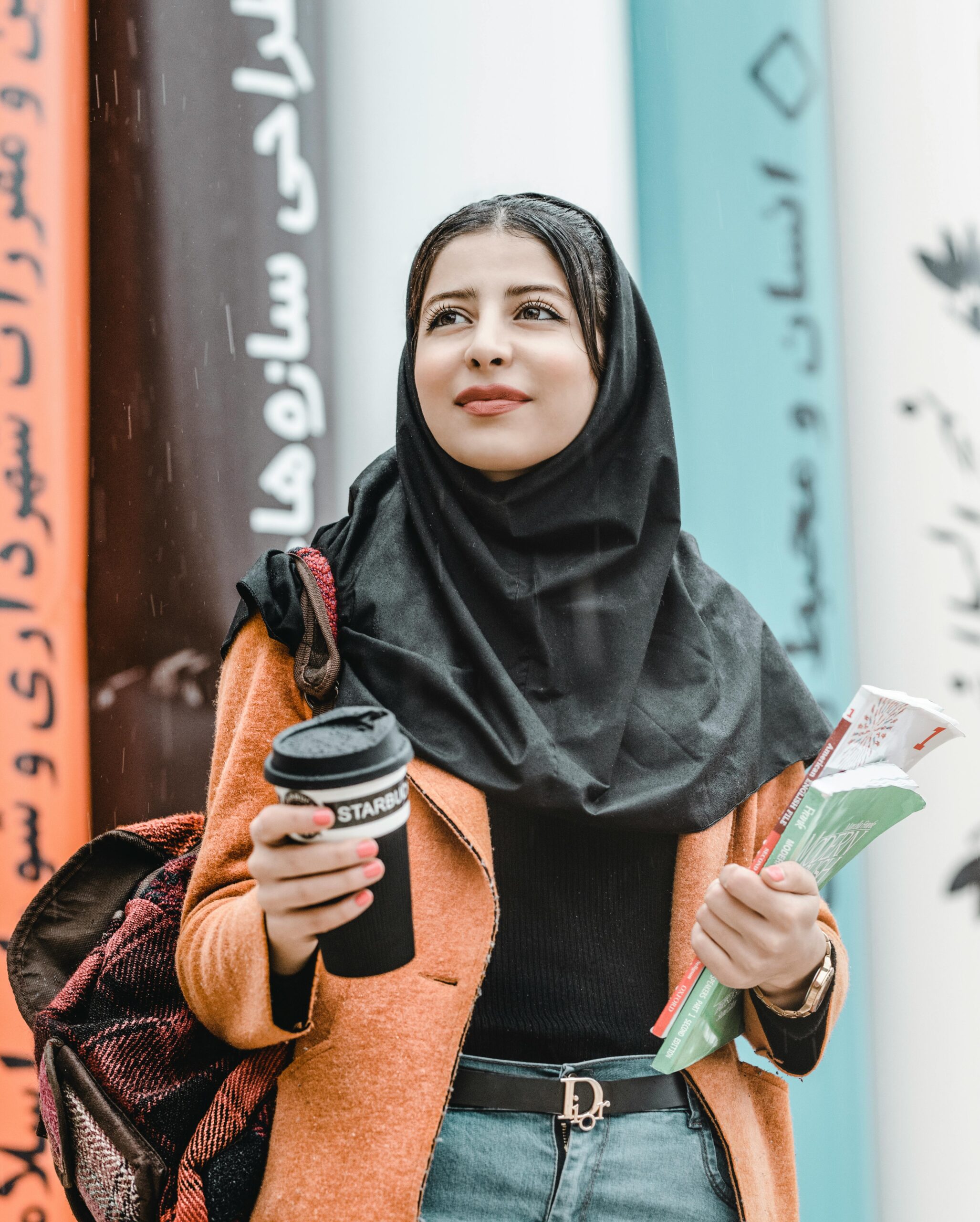 Smiling woman in a hijab holding a coffee cup and books outdoors, showcasing modern fashion and lifestyle.